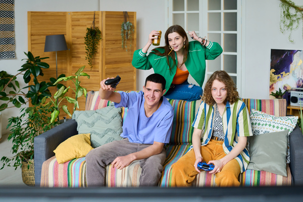 Two young people in bright modern outfits in a bright room with plants are playing a console holding a gamepad in their hands sitting on the sofa on the back of which their girlfriend is sitting with a snack and cheering for victory